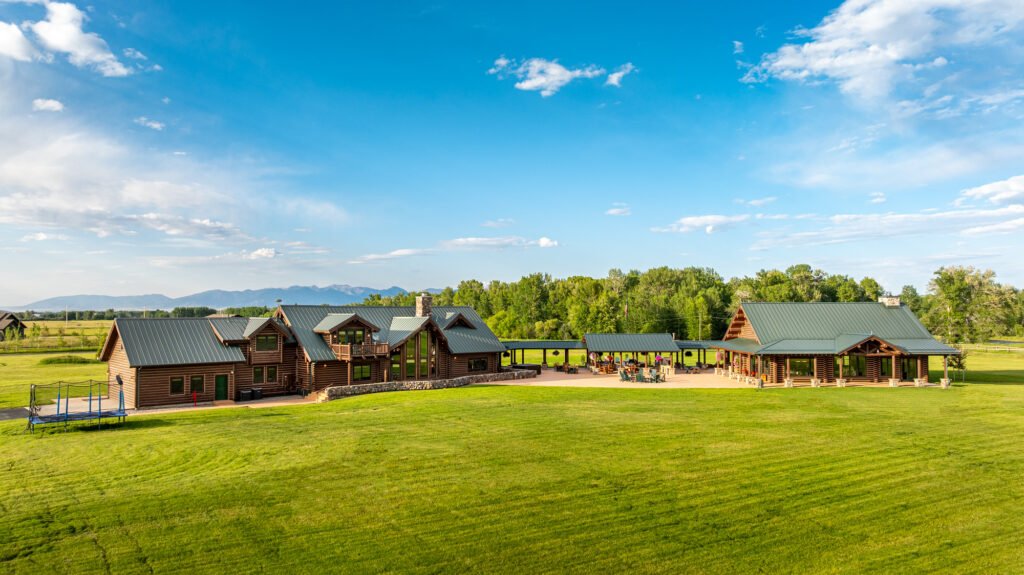 A wide exterior view of the 6,612-SF main log home and expansive gathering spaces at A River’s Ranch in Gallatin Gateway, Montana, featuring a large green lawn, connecting covered breezeways, and mountains in the distance under a blue sky.
