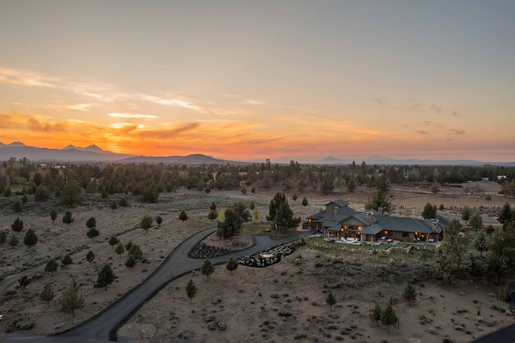 An aerial sunset view of the 2.61-acre custom estate at 61465 White Peaks Court in Bend, Oregon, showing the sprawling multigenerational home and surrounding plains with sweeping views of the distant mountains.