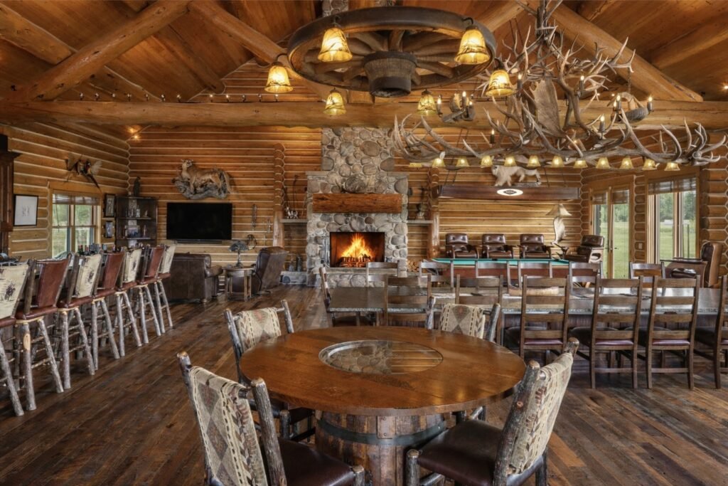 The luxury rustic game room and entertainment space inside the main log home at A River’s Ranch in Gallatin Gateway, Montana, featuring a stone fireplace, large bar counter, dining tables, a pool table, and unique antler and wagon wheel chandeliers.
