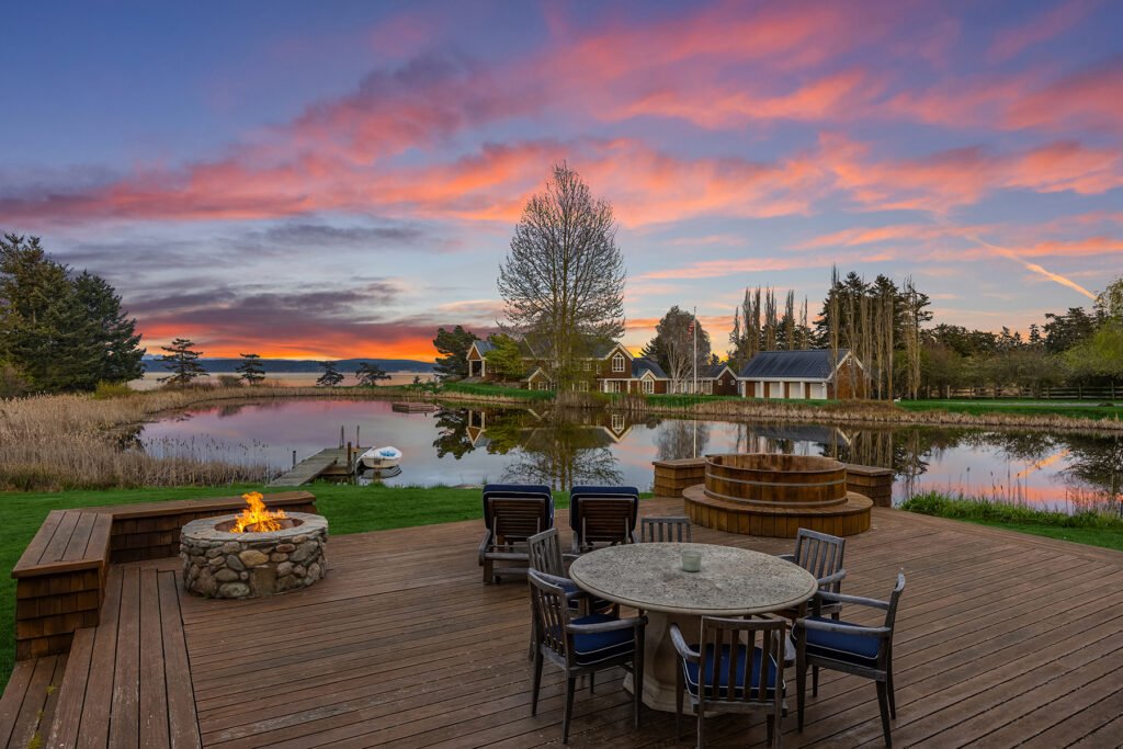 An outdoor wooden entertainment deck at Halftide Farms on San Juan Island, Washington, featuring a stone fire pit, dining table, hot tub, and a scenic view of a private pond and mountains under a vibrant sunset sky.