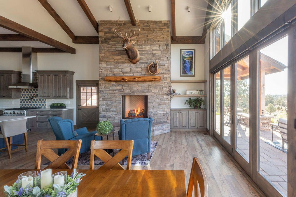 The central open great room at 61465 White Peaks Court in Bend, Oregon, featuring massive vaulted ceilings with custom wood beam accents, a floor-to-ceiling stone fireplace, and large sliding glass doors offering sweeping views of the surrounding plains.