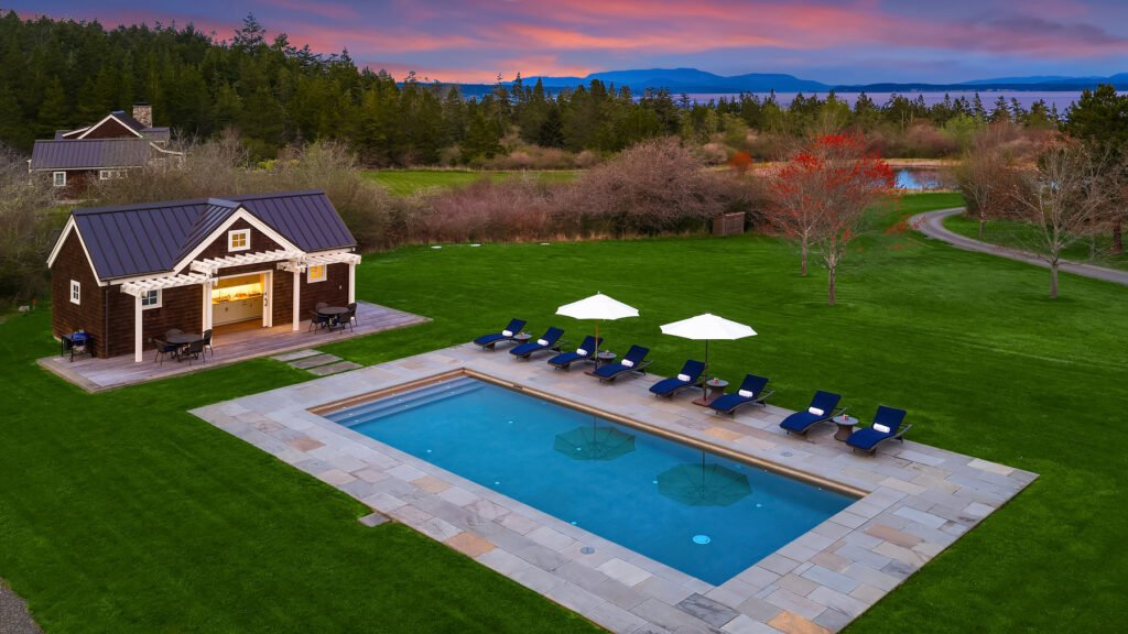 An aerial view of the luxury in-ground swimming pool and adjacent pool house at Halftide Farms on San Juan Island, Washington, featuring a stone patio, lounge chairs, and a vast green lawn with distant mountain and ocean views at sunset.