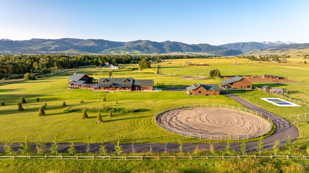 An aerial view of A River’s Ranch at 1021 Gateway South Road in Gallatin Gateway, Montana, showcasing the 6,612-SF main log home, a private guest house, extensive equestrian facilities with an arena, and expansive usable acreage set against distant mountain ranges.