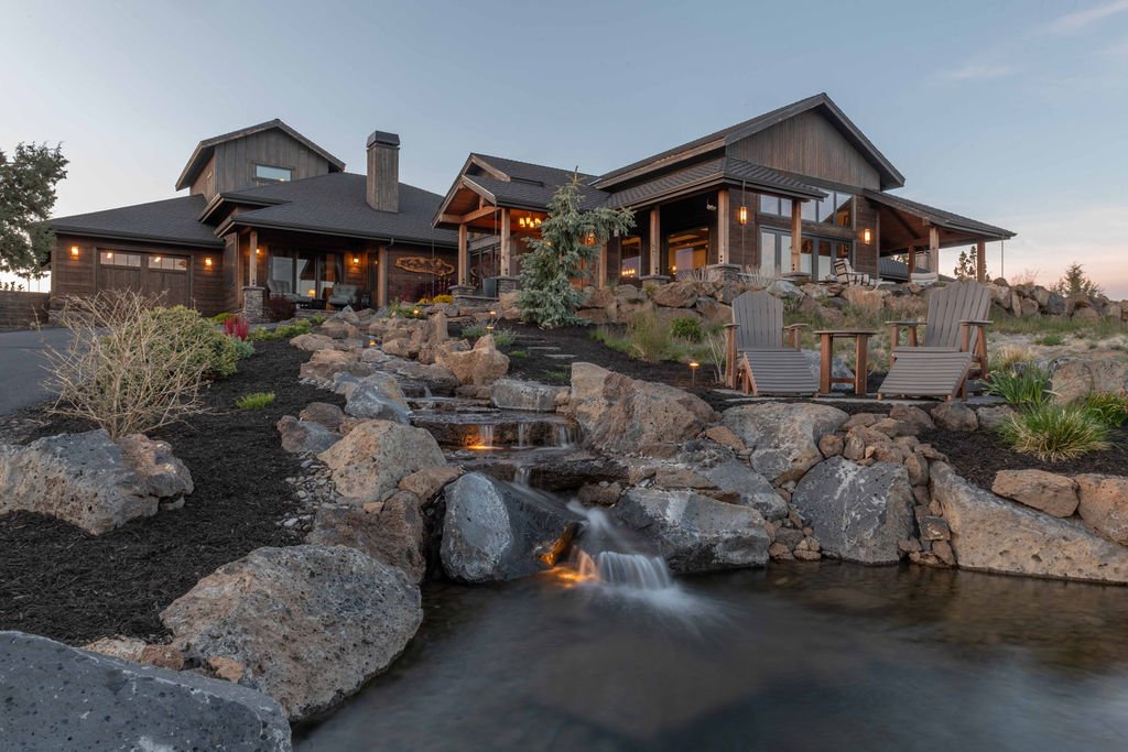 A front exterior view of the custom multigenerational estate at 61465 White Peaks Court in Bend, Oregon, featuring a luxury modern rustic home design, natural stone cascading water feature, and outdoor lounge seating at dusk.