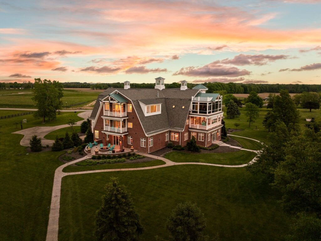 An aerial sunset view of the massive 25,000-square-foot waterfront estate at 9825 Windy Hills Drive in Zionsville, Indiana, showing the multi-story luxury home, stone patio with a fire pit, and expansive green grounds under a vibrant sky.