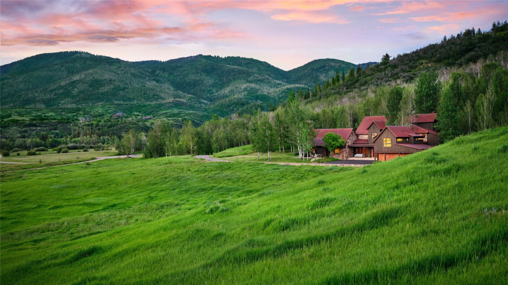 An aerial view of the Meadow House luxury mountain retreat at Storm Mountain Ranch in Steamboat Springs, Colorado, nestled between open green ranch meadows and dense aspen groves against rolling green hills at sunset.