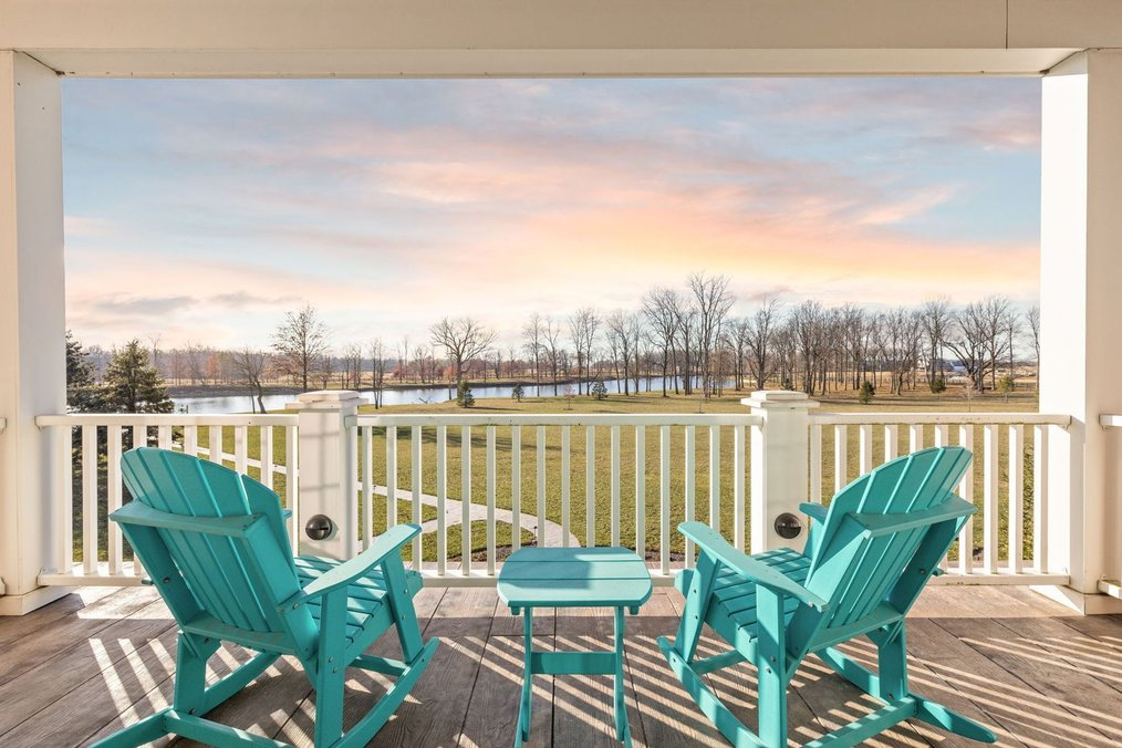 A covered upper deck view at 9825 Windy Hills Drive in Zionsville, Indiana, featuring two bright blue Adirondack rocking chairs overlooking a vast green lawn, bare winter trees, and a 35-acre lake at sunset.