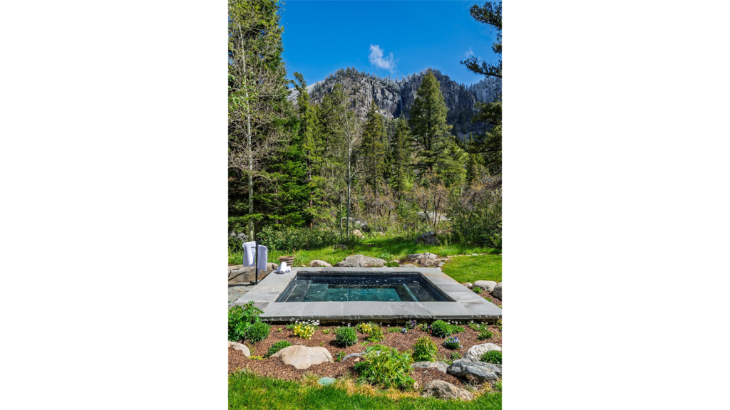 An outdoor stone hot tub at Storm Mountain Ranch in Steamboat Springs, Colorado, surrounded by lush green landscaping, pine trees, and a dramatic mountain ridge with a visible waterfall under a clear blue sky.