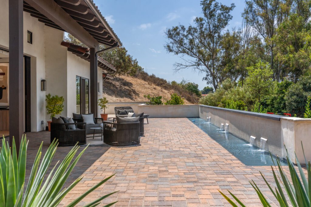 The spacious outdoor stone-paved patio at 5758 Las Palomas in Rancho Santa Fe, California, featuring a built-in water feature with cascading fountains, a contemporary outdoor lounge seating area, and white stucco walls under a sunny sky.