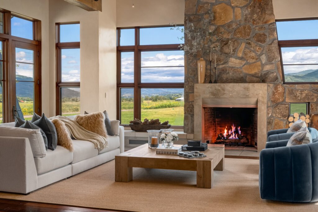 The luxury mountain living room interior at Meadow House in Storm Mountain Ranch, Steamboat Springs, Colorado, featuring a floor-to-ceiling stone fireplace, modern white sectional sofa, blue accent chairs, a wooden coffee table, and large windows framing scenic valley views.