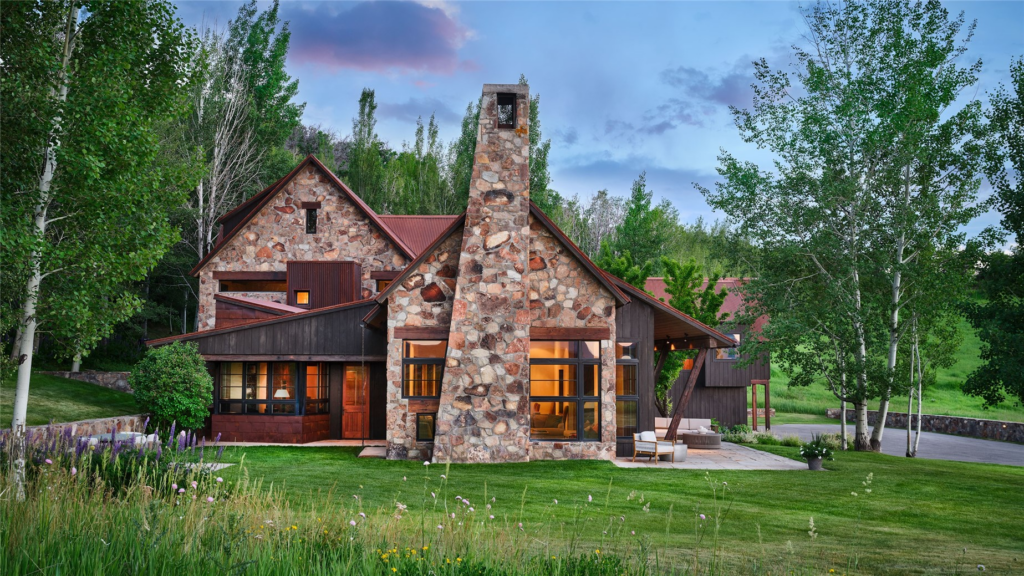 A front exterior view of the Meadow House at Storm Mountain Ranch in Steamboat Springs, Colorado, designed by architect Michael Ernemann, featuring a custom natural stone chimney, wood siding, and warm indoor lighting surrounded by green grass and aspen trees.