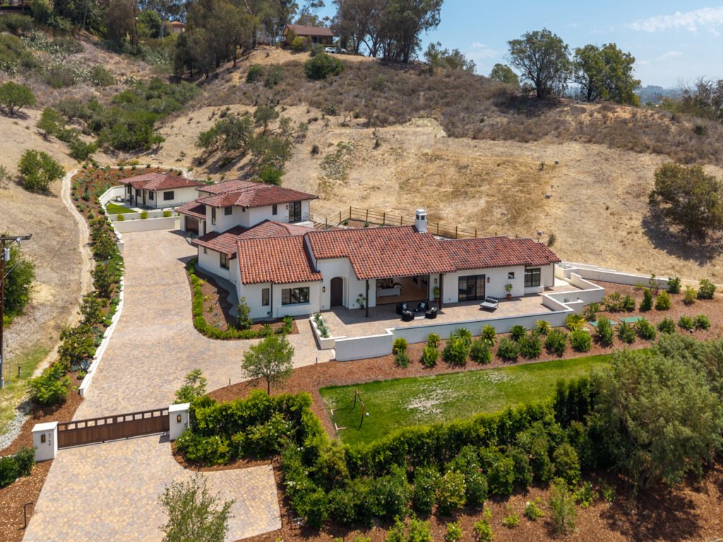 An aerial view of the newly constructed modern Santa Barbara style estate at 5758 Las Palomas in Rancho Santa Fe, California, featuring a multi-generational compound with red tile roofs, white stucco walls, private gates, a detached guest house, and a paved driveway nestled against a hillside.