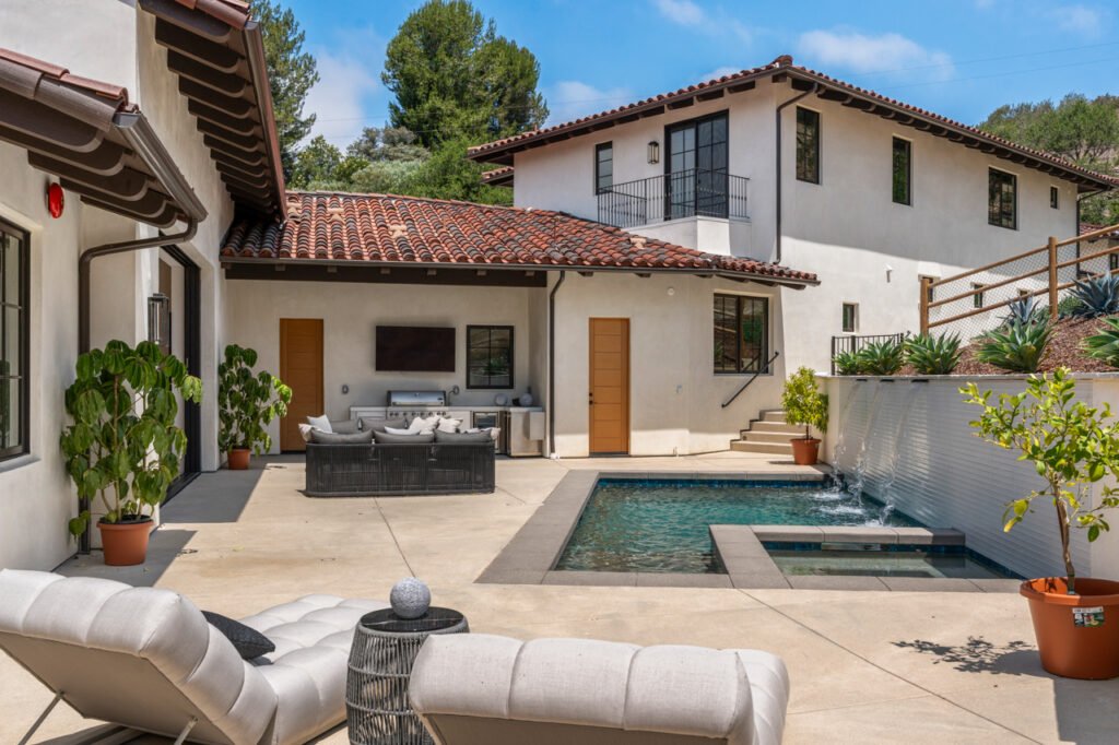 The luxury outdoor pool patio and built-in kitchen area at 5758 Las Palomas in Rancho Santa Fe, California, featuring an in-ground swimming pool with a cascading water wall, lounge chairs, and a detached guest house structure in the background.