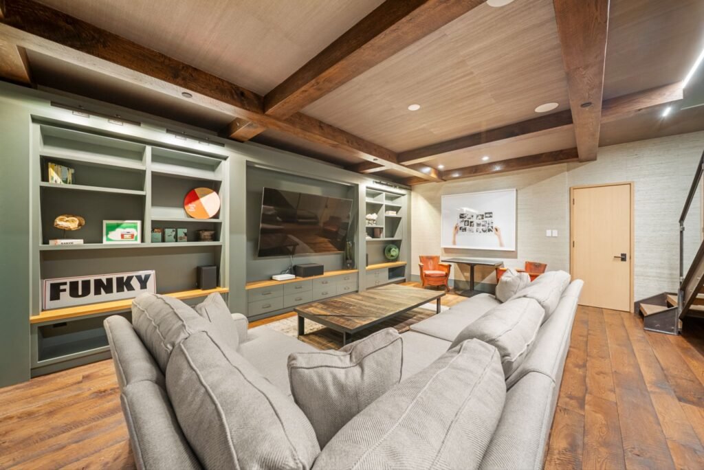 Luxury lower-level media room at 150 W Pacific Avenue in Telluride, featuring a large grey sectional couch, sage green custom built-in entertainment shelving, a rustic wood-coffered ceiling, and pop art decor.