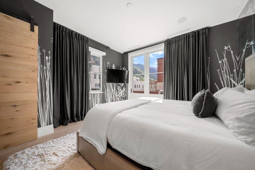 Contemporary bedroom interior at 150 W Pacific Avenue in Telluride, featuring custom dark wallpaper with white botanical outlines, a sliding wood barn door, full-length charcoal curtains, and windows with mountain views.