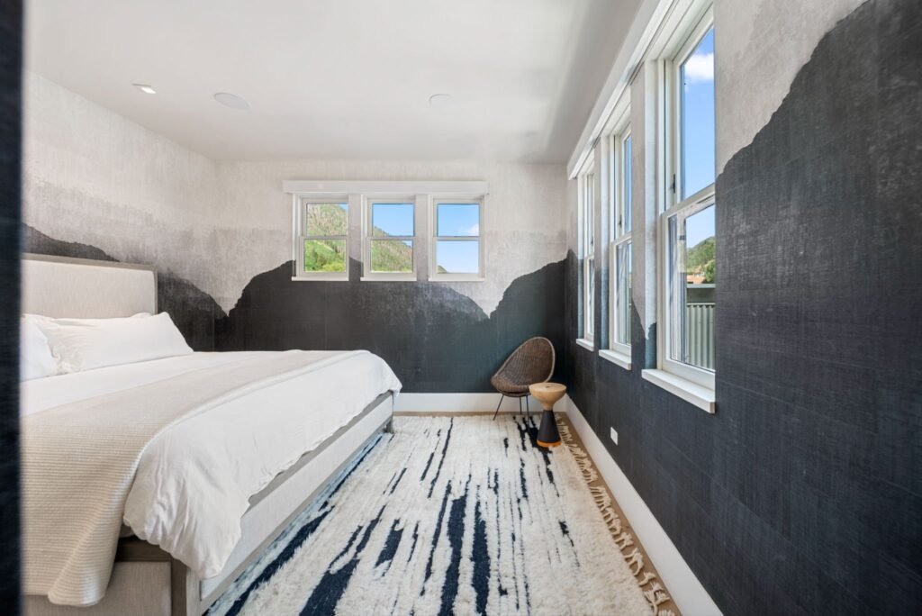 Modern guest bedroom at 150 W Pacific Avenue in Telluride, featuring custom abstract mountain-silhouette wallpaper, a plush white bed, a textured area rug, and large windows looking out toward downtown.