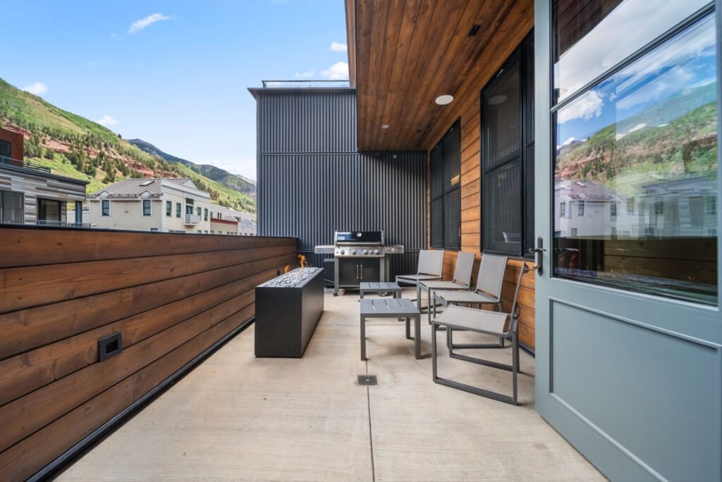 Modern outdoor entertainment patio at 150 W Pacific Avenue in Telluride, featuring a wood-planked balcony with a built-in stainless steel grill, a contemporary rectangular fire pit, a metal lounge seating set, and stunning views of the surrounding Rocky Mountain peaks.