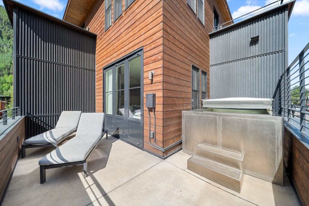 Luxury modern mountain patio at 150 W Pacific Avenue in Telluride, featuring a custom built-in metal-sided jacuzzi hot tub, two comfortable lounge chairs, and an exterior of rich wood paneling mixed with dark corrugated metal accents.