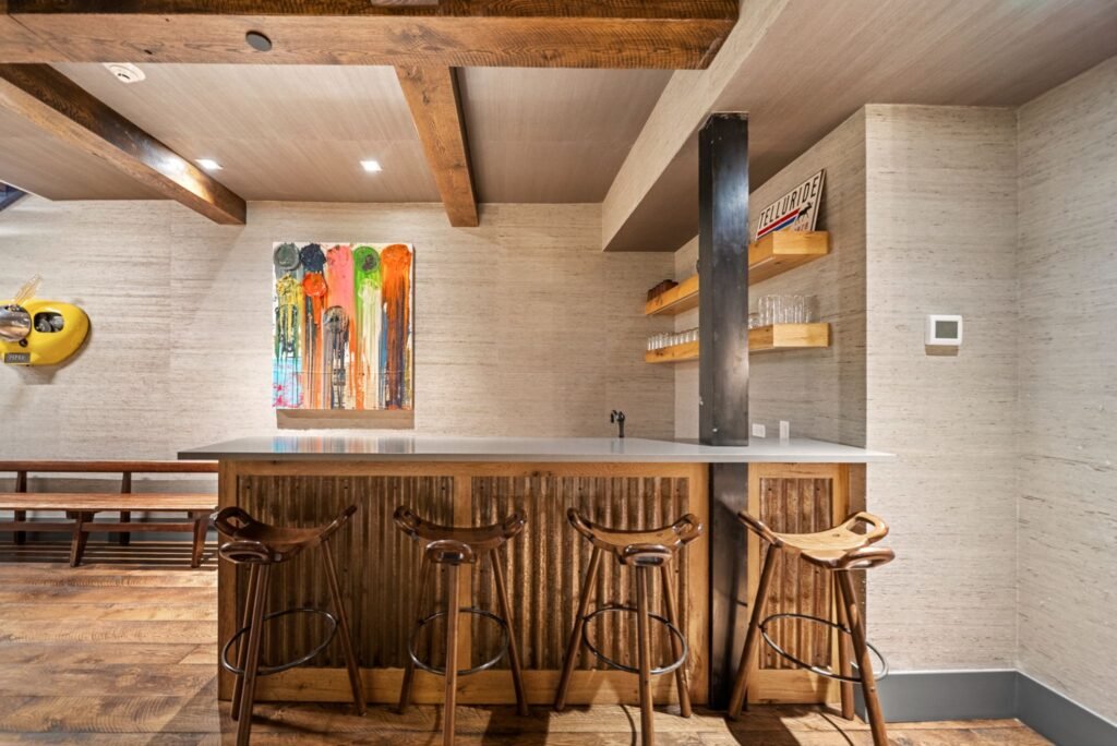 Rustic modern home bar lounge at 150 W Pacific Avenue in Telluride, featuring a corrugated metal bar front, wooden barstools, floating shelves, exposed ceiling beams, and colorful contemporary artwork.