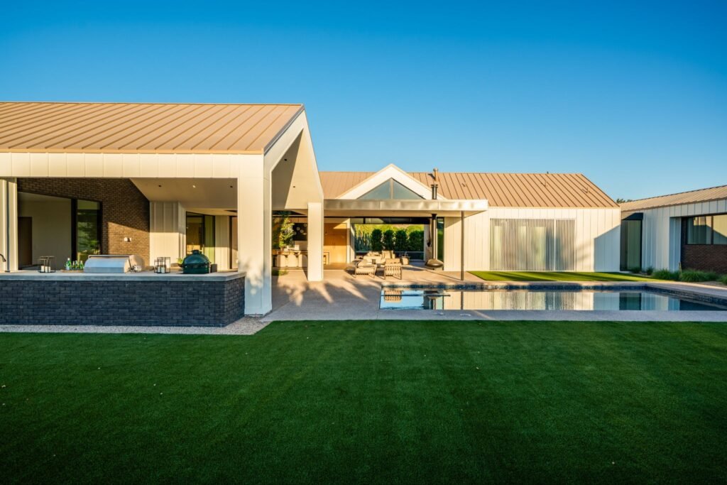 Modern desert home architecture by The Ranch Mine in Paradise Valley, AZ, showcasing a metal gable roof line, vertical white siding, an outdoor brick kitchen bar, and a luxury swimming pool layout.