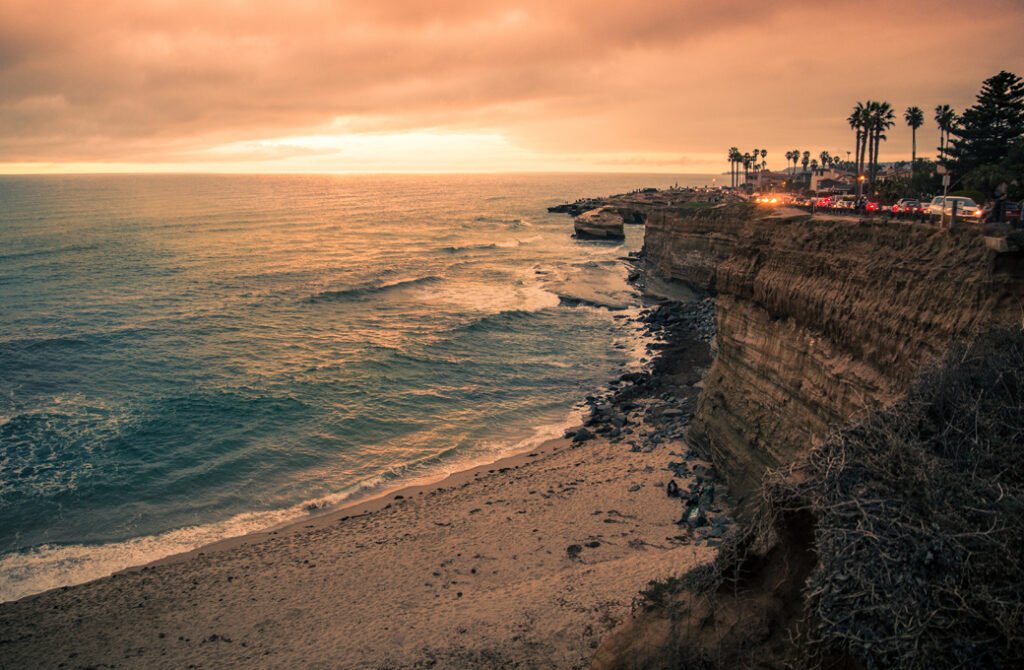 Scenic golden hour sunset over the ocean at Sunset Cliffs in San Diego, California, featuring rugged coastline cliffs, a sandy beach, and palm trees.