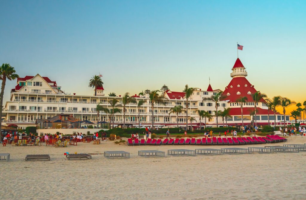 The iconic Hotel del Coronado beach resort in San Diego, California, featuring its historic red-roofed Victorian building and rows of lounge chairs on the sand during sunset.