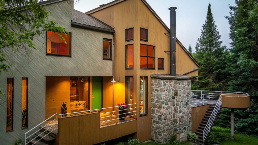 Dusk architectural exterior of 320 Ch Graham in Ivry-sur-le-Lac, Quebec featuring warm glowing interior lights, cedar wood paneling, a stone chimney, and an elevated wooden deck surrounded by trees.
