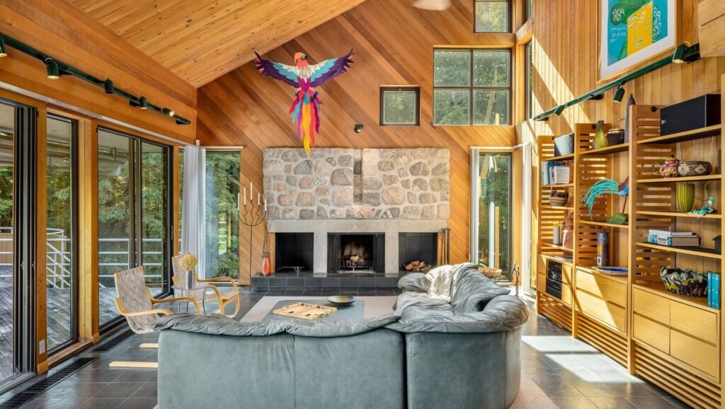 Luxury living room interior at 320 Ch Graham in Ivry-sur-le-Lac, Quebec featuring soaring cedar cathedral ceilings, a massive stone fireplace, a grey leather sectional sofa, and bright floor-to-ceiling windows.