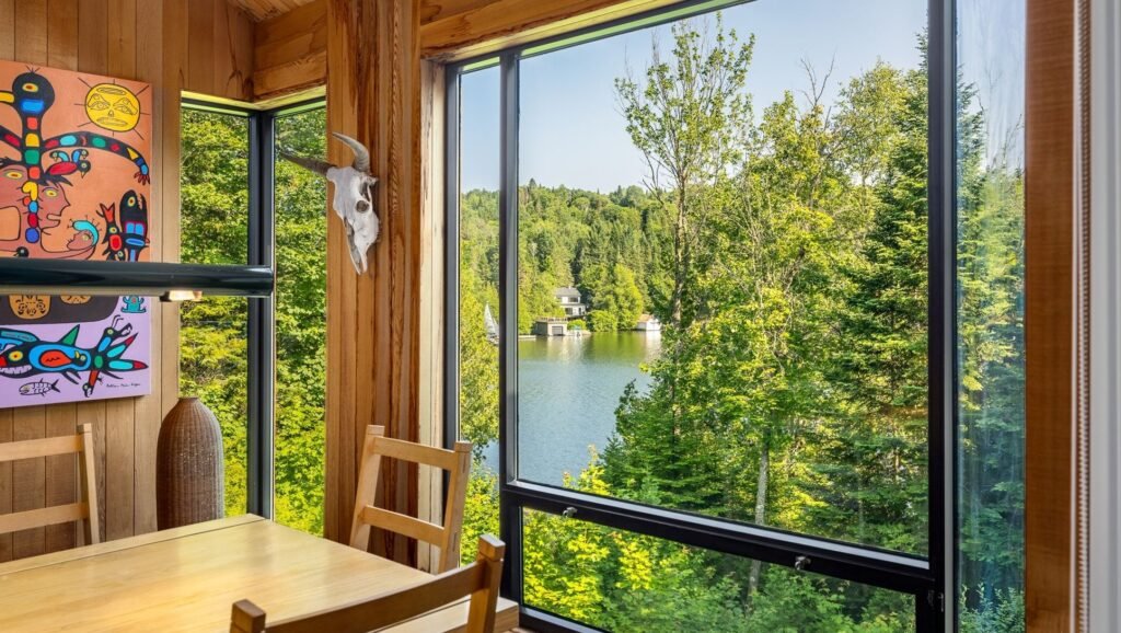 Rustic modern dining room interior at 320 Ch Graham in Ivry-sur-le-Lac, Quebec featuring natural wood walls, colorful Indigenous artwork, and a large window framing scenic views of Lac Manitou.