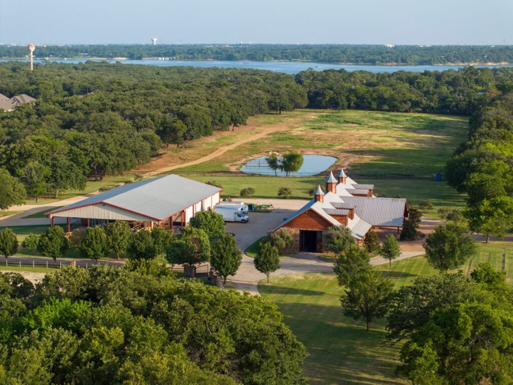 An aerial view of the equestrian facilities and barns on the 50-acre legacy estate outside Dallas, Texas, showing a private pond, open pastures, and mature trees with a lake visible in the distance.