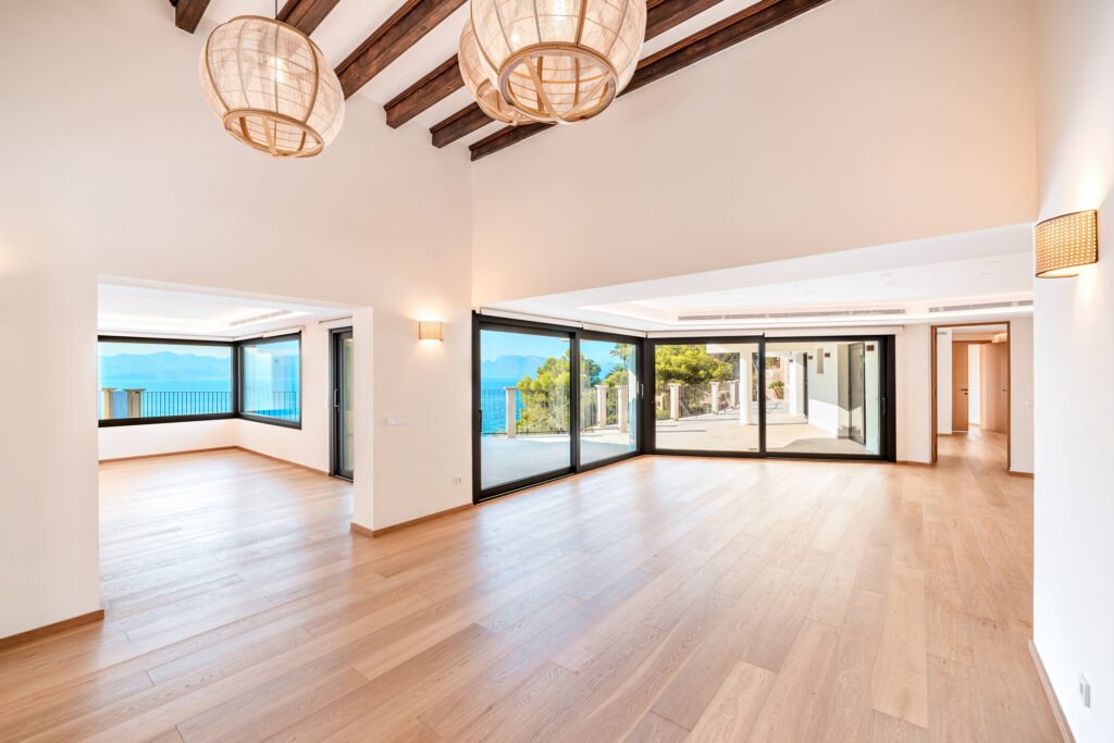 Modern open-plan living room with exposed wood beams, hardwood flooring, and large sea-view glass doors in a luxury Mallorca villa.
