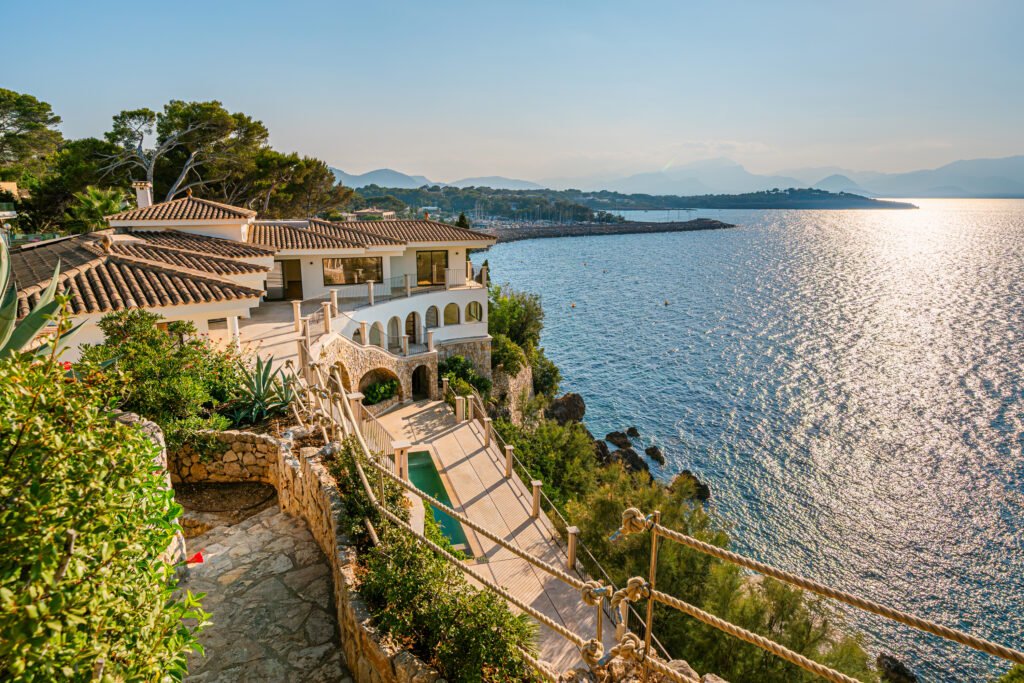 High-angle view of a cliffside luxury villa with terracotta tile roofs, a swimming pool terrace, and panoramic sea views in Bonaire, Mallorca.