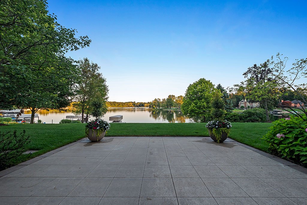 Outdoor stone patio and manicured green lawn at 4969 Elmgate Drive, Orchard Lake, Michigan, showcasing a scenic lakeside view, vibrant flower planters, and the calm waters of Upper Straits Lake.