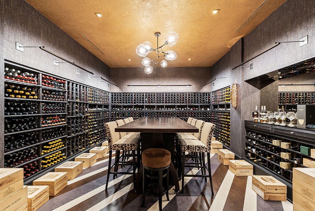 State-of-the-art custom wine cellar and tasting room at 4969 Elmgate Drive, Orchard Lake, Michigan, featuring extensive floor-to-ceiling storage racks, a central tasting table with upholstered barstools, a modern glass globe chandelier, and a dedicated sommelier service bar.