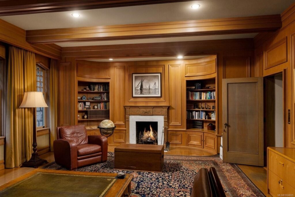 Wood-paneled library and study inside the historic Riffington estate at 3175 Beach Drive, Oak Bay, British Columbia, featuring custom built-in bookshelves, an ornate fireplace mantel, exposed ceiling beams, and a leather armchair.