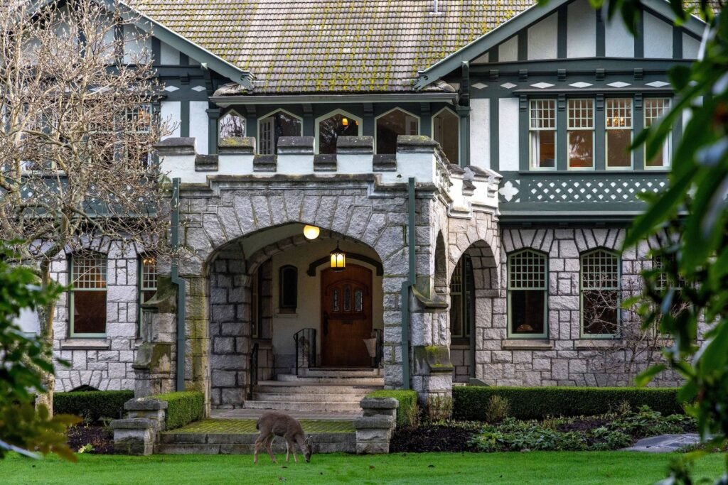 Historic stone portico entrance of the Riffington estate at 3175 Beach Drive, Oak Bay, British Columbia, showcasing a grand granite archway, Tudor Revival timber details, a multi-pane grid window layout, and a wild deer grazing on the manicured front lawn.
