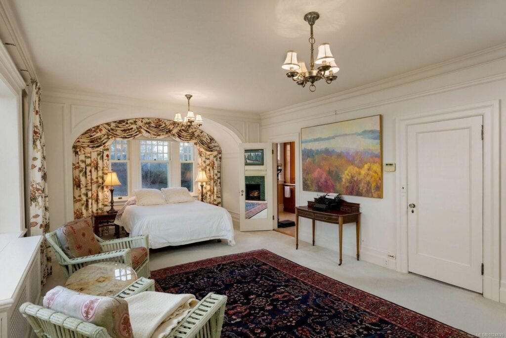 Elegant luxury bedroom suite at the historic Riffington estate at 3175 Beach Drive, Oak Bay, British Columbia, featuring a custom arched bed alcove with floral drapery, an antique writing desk, traditional chandeliers, and a large oriental rug.