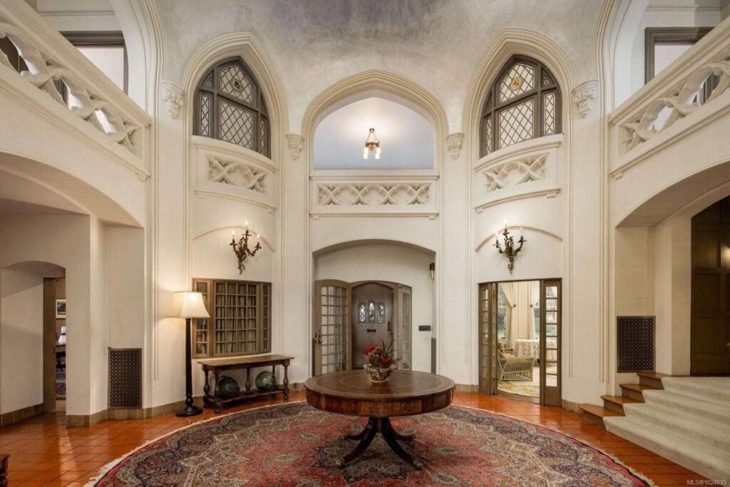 Two-story reception hall at the historic Riffington estate at 3175 Beach Drive, Oak Bay, British Columbia, featuring a double-height rotunda, Gothic-style arched windows, stone-look architectural moldings, a central round table, and terracotta tile flooring.