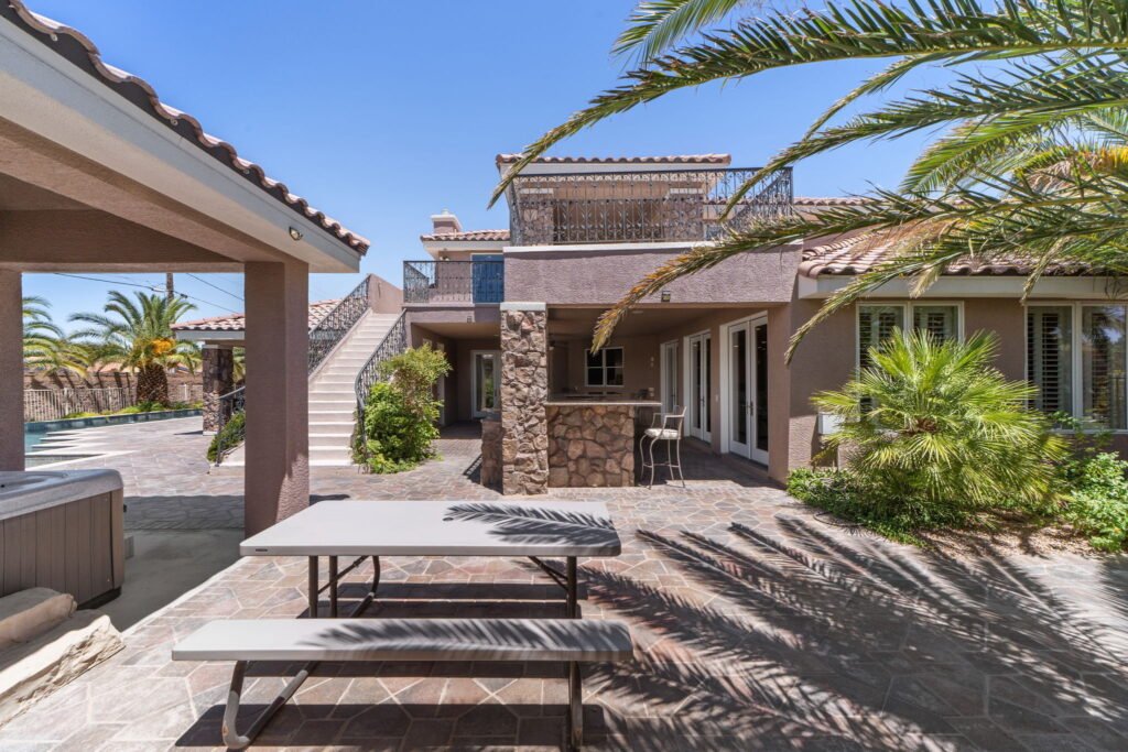 Rear courtyard and outdoor entertainment space at 208 E Levi Avenue in Las Vegas showcasing a custom stone-clad bar, an upper-level balcony terrace with a wrought-iron railing, a patio picnic table, and palm tree landscaping.