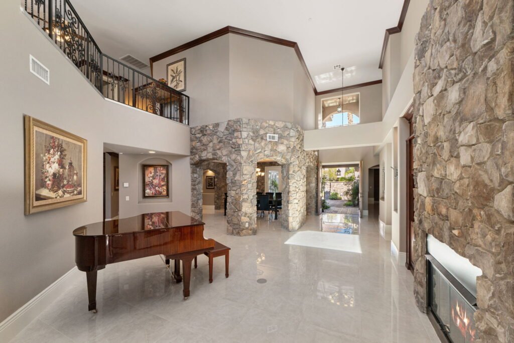 Foyer interior of a custom mansion at 208 E Levi Avenue in Las Vegas highlighting high ceilings, an open entryway, a polished wood grand piano, a stone fireplace, and a second-story landing with wrought-iron railings.