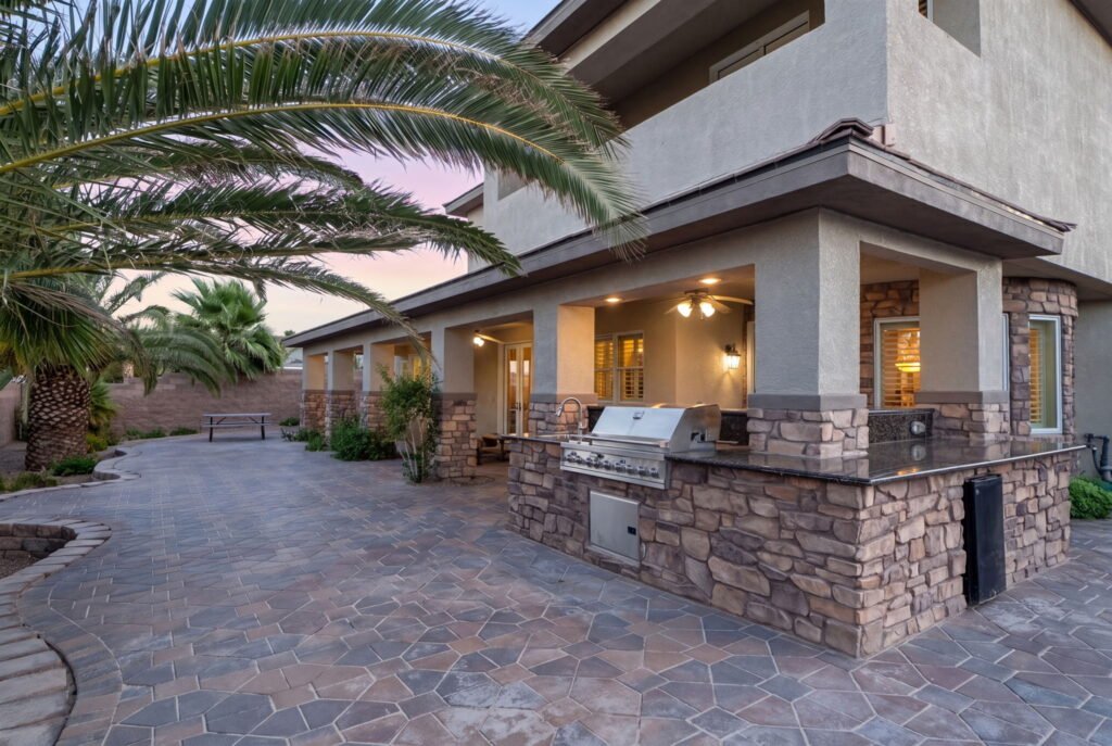 Covered patio and outdoor kitchen setup at 208 E Levi Avenue in Las Vegas highlighting a built-in stone BBQ island, granite countertops, a flagstone paved deck, and a shaded backyard living area.