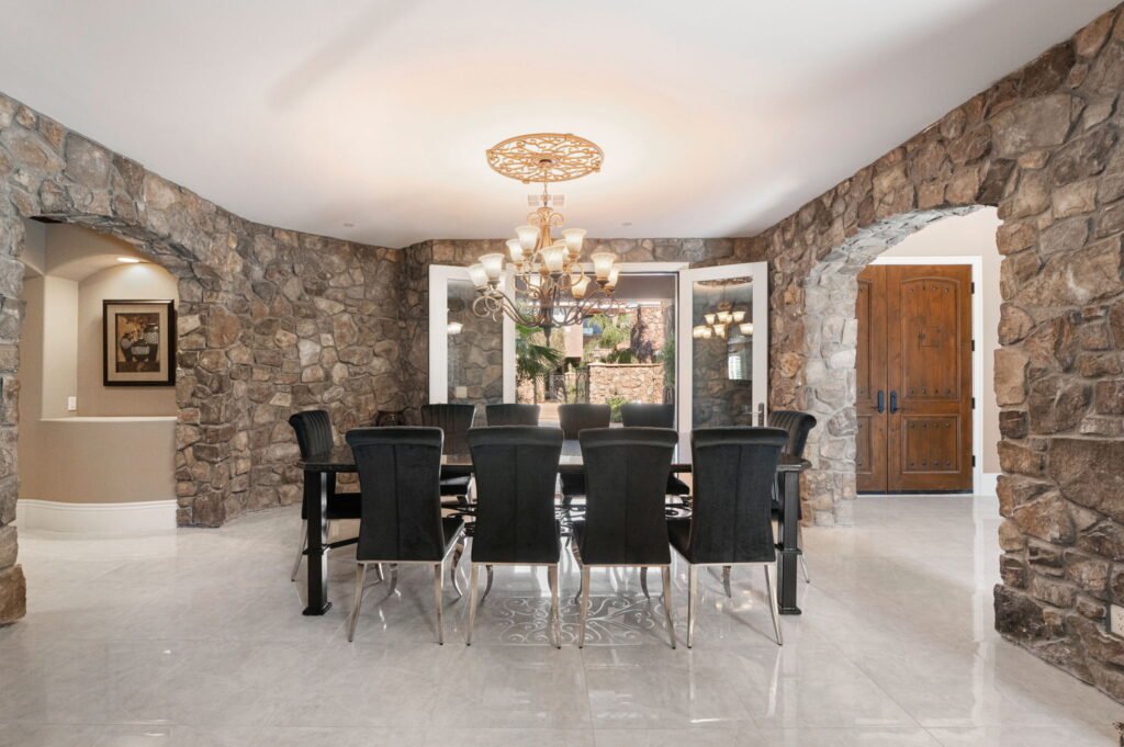 Formal dining space inside a custom home at 208 E Levi Avenue in Las Vegas showing stone accent walls, stone archways, velvet high-back chairs, polished marble flooring, and double entry doors.