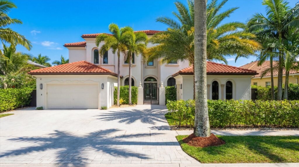 Front exterior view of the Mediterranean-style estate at 2186 Stotesbury Way in the Olympia community of Wellington, Florida, featuring a two-story light stucco facade, terracotta tile roofing, a wide paver driveway with an attached garage, a secure gated courtyard entry, and a beautifully landscaped yard lined with mature palm trees.