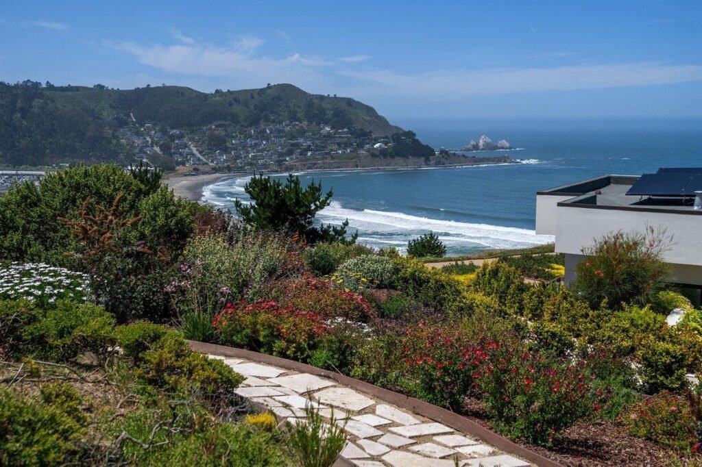 Panoramic ocean and coastline view from the gated estate at 5 Ohlone Drive in Pacifica, California, showcasing rolling ocean waves, a crescent beach, a stone walking path, lush hillside gardens, and the distant coastal mountains of the Bay Area under a bright blue sky.