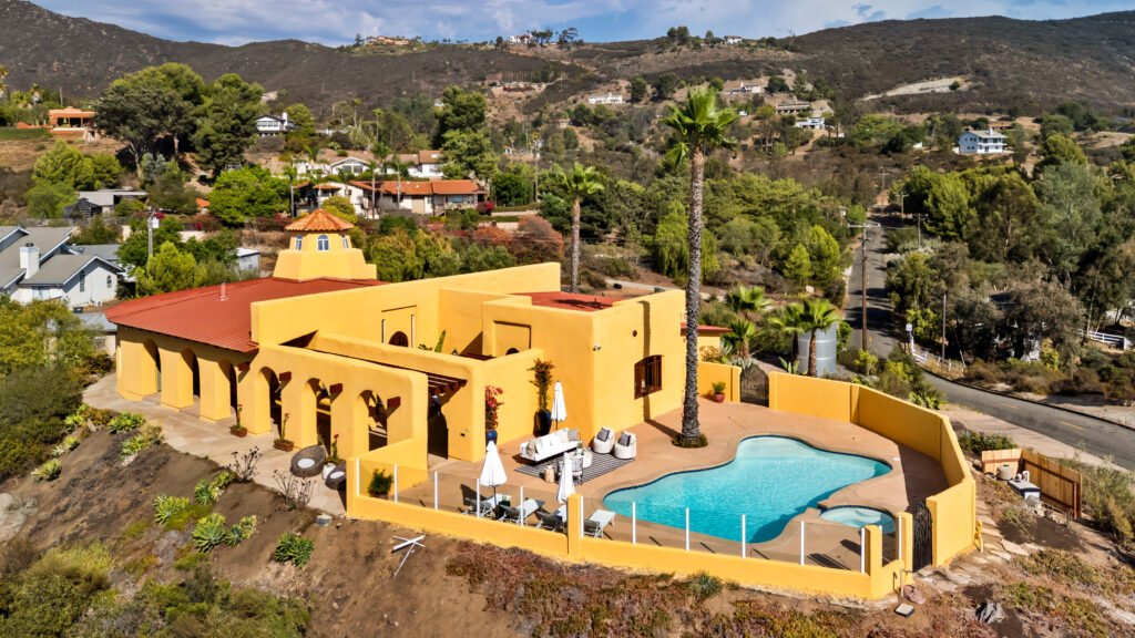 Aerial view of the unique Spanish-style estate at 3696 Alta Loma Drive in Jamul, California, highlighting its bright yellow stucco facade, terracotta tile roof with a prominent dome tower, a curved swimming pool and spa terrace, and its private hillside position in rural San Diego County.