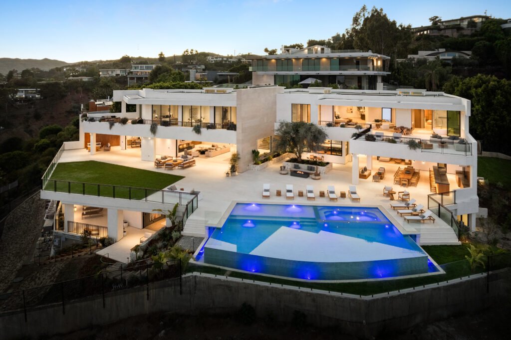 Twilight aerial view of the contemporary hillside mega-mansion at 1204 Chickory Lane in Brentwood, Los Angeles, highlighting its white tiered architectural facade, illuminated infinity pool and integrated spa, expansive lounge terraces, manicured lawn, and panoramic canyon vistas.