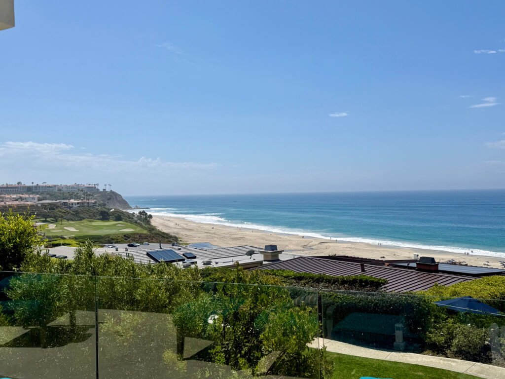 Panoramic ocean and coastline view from the off-market luxury estate at 416 Monarch Bay Drive in Dana Point, California, looking past a glass balcony railing over coastal rooftops toward Salt Creek Beach and the Pacific Ocean under a bright blue sky.