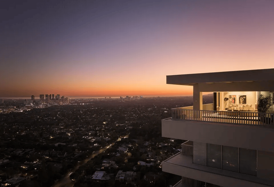 Sunset view from the luxury penthouse balcony at 9255 Doheny Road in the iconic Sierra Towers of West Hollywood, California, looking out over the illuminated Los Angeles city skyline and distant skyscrapers under a gradient orange and purple evening sky.