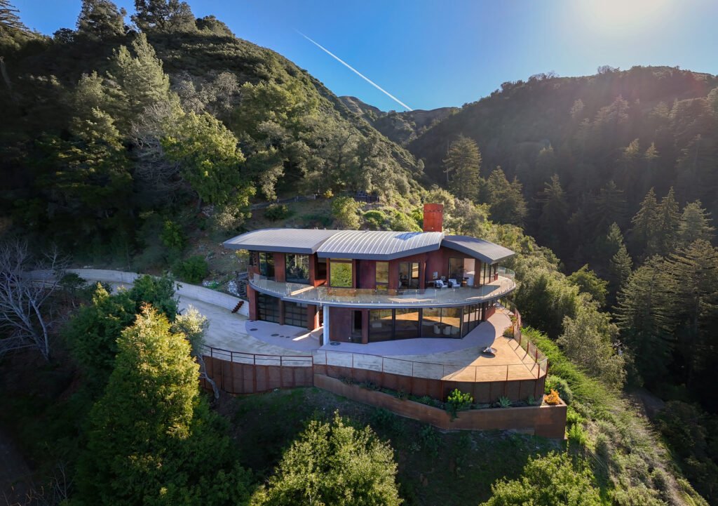 Aerial view of the modern architectural home at 47730 Coast Ridge Road in Big Sur, California, showcasing its curved zinc-colored roof, sweeping glass balconies, multi-level design, and secluded hillside placement surrounded by dense redwood forests and coastal mountains.