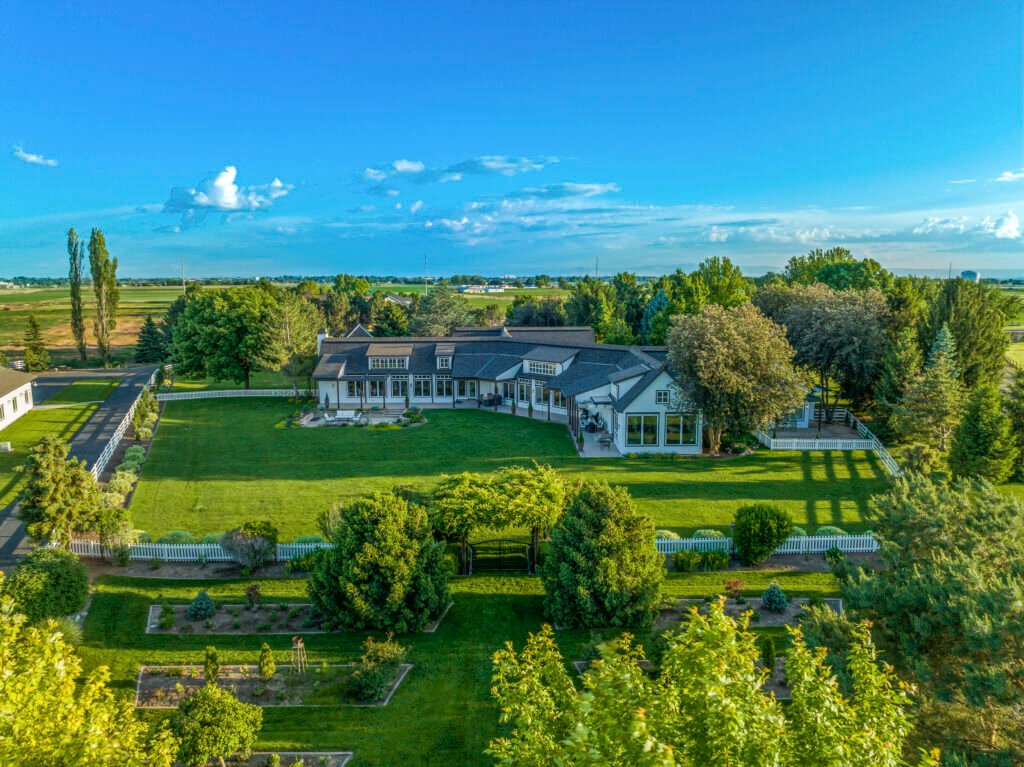 Elevated aerial view of the expansive equestrian estate at 7351 West Ledgerwood Lane in Meridian, Idaho, showcasing the multi-wing 6,451-square-foot main residence, a sprawling green lawn enclosed by white picket fencing, manicured gardens, and mature trees under a vibrant blue sky.